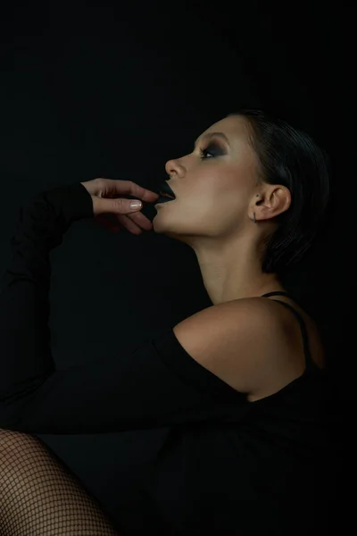 Готический гламур, профиль очаровательной женщины с жутким макияжем, касающимся темных губ на черном фоне — стоковое фото