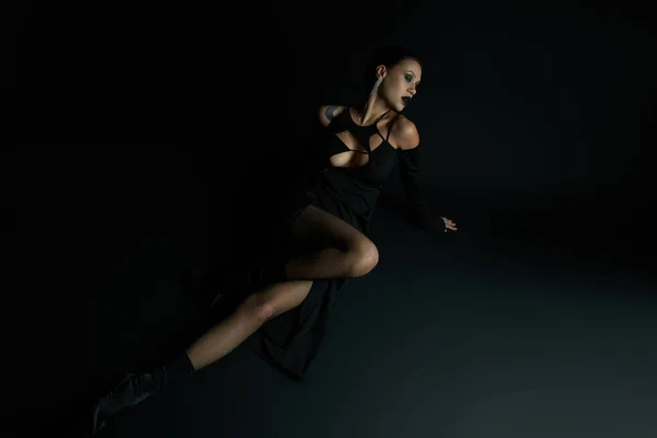Mujer inspirada en el demonio tatuado en vestido de halloween elegante sentado sobre fondo negro, longitud completa - foto de stock