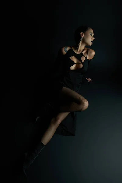 Geheimnisvolle tätowierte Frau in sexy Halloween-Kleid posiert mit geschlossenen Augen, während sie auf schwarz sitzt — Stockfoto