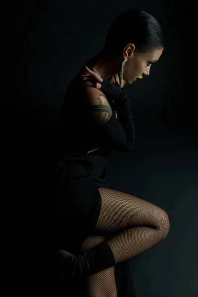 Side view of alluring tattooed woman in sexy dress and fishnet tights sitting on black, dark beauty — Stock Photo