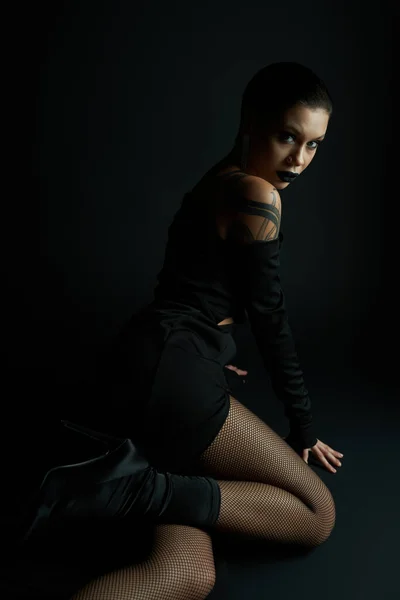 Leidenschaftliche Frau in sexy Kleid und Netzstrumpfhose sitzt und schaut in die Kamera vor schwarzem Hintergrund — Stockfoto
