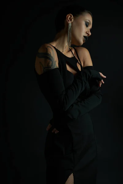 Татуированная женщина в стильном платье на Хэллоуин и темный макияж позирует с закрытыми глазами на черном фоне — стоковое фото