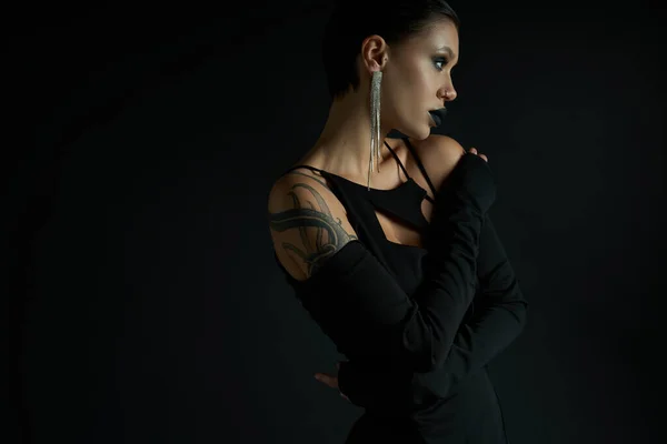 Tätowierte und glamouröse Frau mit schaurigem Make-up posiert in sexy Kleid auf schwarzem, halloween-artigen Konzept — Stockfoto