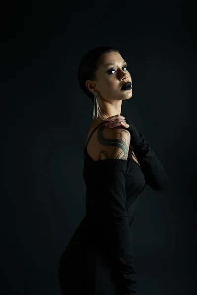 Femme tatouée élégante avec des lèvres sombres portant une robe d'Halloween sexy et regardant la caméra sur noir — Photo de stock