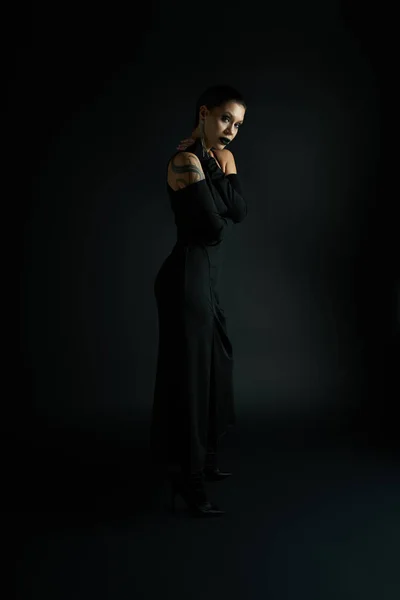 Sexy tatuado mulher com escuro lábios vestindo elegante halloween vestido e olhando para câmera no preto — Fotografia de Stock