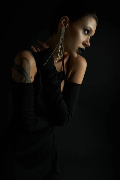 Retrato de mujer tatuada seductora en vestido de halloween elegante y sexy mirando hacia otro lado en negro - foto de stock
