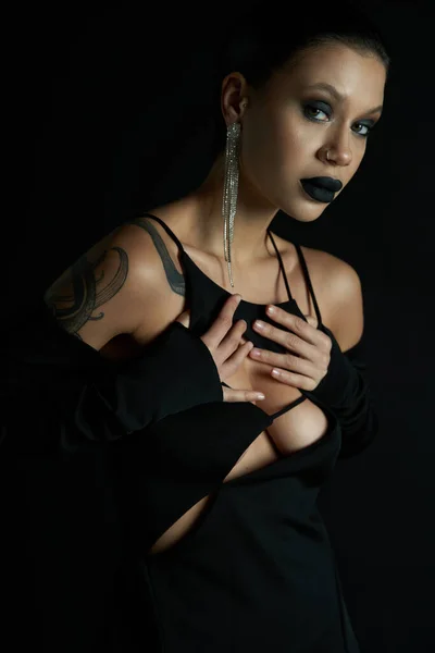 Encanto gótico, mujer tatuada en vestido de halloween sexy tocando el pecho y mirando a la cámara en negro - foto de stock