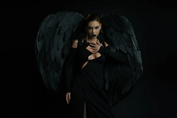 Сексуальная женщина в костюме падшего ангела с темными крыльями, смотрящая в камеру на черном, концепция Хэллоуина — стоковое фото