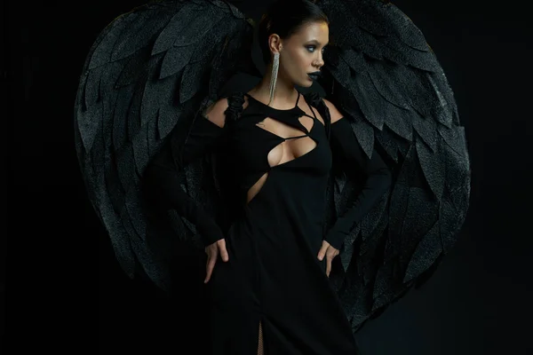 Mujer en maquillaje oscuro y traje de fantasía de criatura alada demoníaca mirando hacia otro lado en negro - foto de stock
