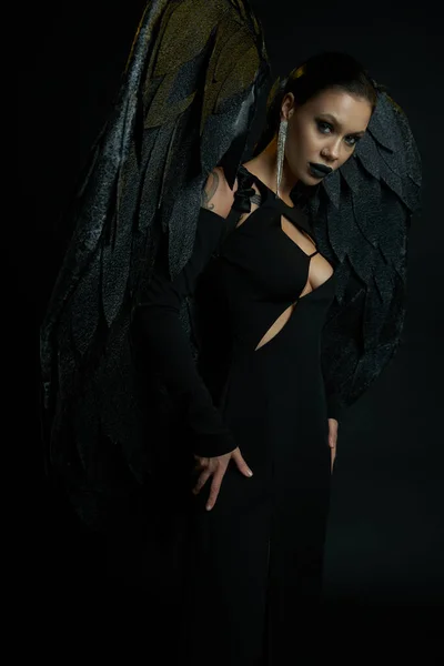 Сексуальная татуированная женщина в костюме падшего ангела на Хэллоуин с темными крыльями, смотрящая в камеру на черном — стоковое фото