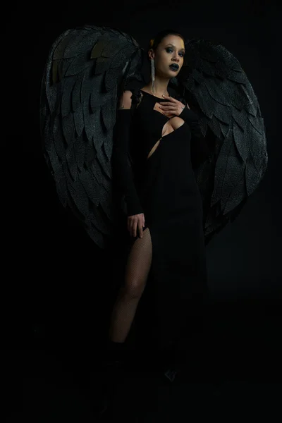 Donna in abito sexy e fantasia ali demoniache scure guardando la fotocamera su nero, concetto di Halloween — Foto stock