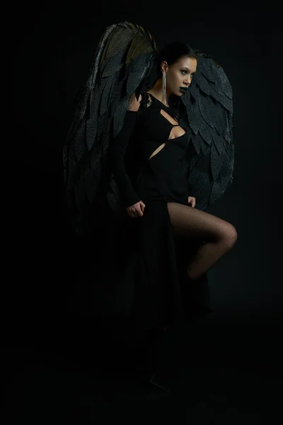 Seductora mujer en maquillaje oscuro y disfraz con alas demoníacas mirando hacia otro lado en negro, Halloween - foto de stock