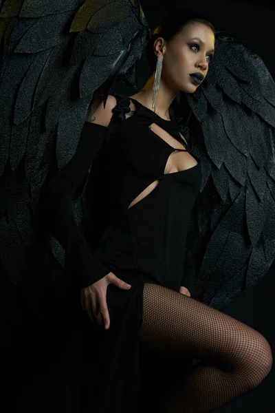 Sexy mujer en halloween traje de demoníaco alado criatura y espeluznante maquillaje mirando hacia otro lado en negro - foto de stock