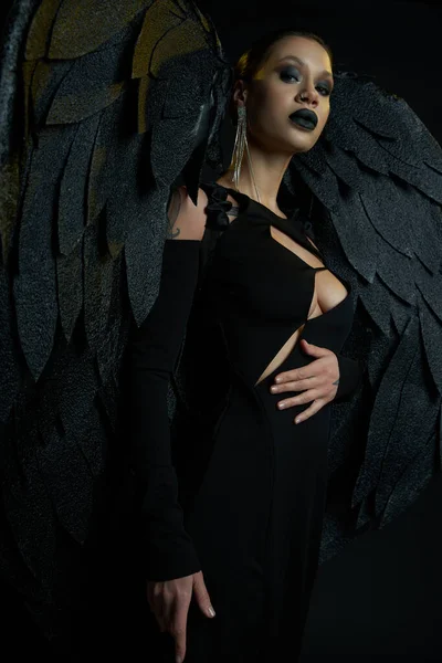 Dark beauty, tattooed woman in halloween costume of winged fallen angel looking at camera on black — Stock Photo