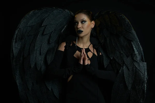 Mujer tatuada en traje de ángel oscuro de pie con las manos de oración y mirando hacia otro lado en negro - foto de stock