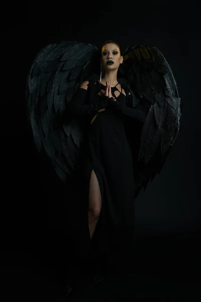Donna misteriosa in costume di creatura alata in piedi con le mani in preghiera su nero, bellezza demoniaca — Foto stock
