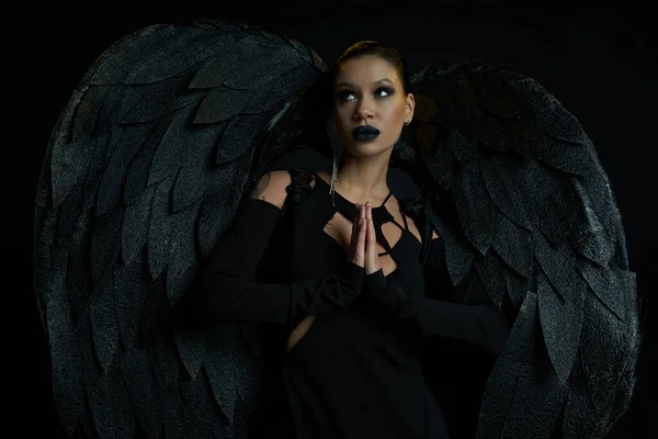 Сексуальная женщина в костюме темного падшего ангела, стоящего с молитвенными руками и смотрящего на черное — стоковое фото