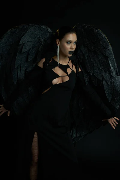 Verführerische Frau im Kostüm eines dunklen Dämons mit Flügeln, die auf schwarzes Halloween-Konzept wegschauen — Stockfoto
