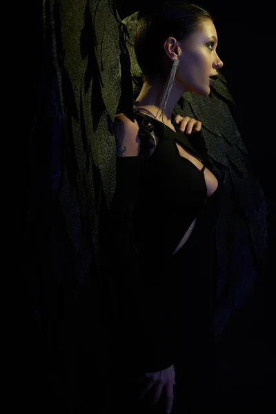 Демоническое обаяние, вид сбоку на сексуальную женщину в Хэллоуинском костюме темного демона с крыльями на черном — стоковое фото