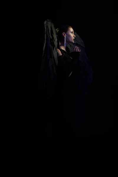 Visão lateral da mulher encantadora como demônio escuro com asas orando em pano de fundo preto, conceito halloween — Fotografia de Stock