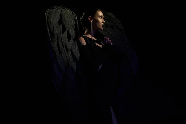 Vista lateral de mujer misteriosa disfrazada de criatura alada demoníaca rezando sobre fondo negro - foto de stock