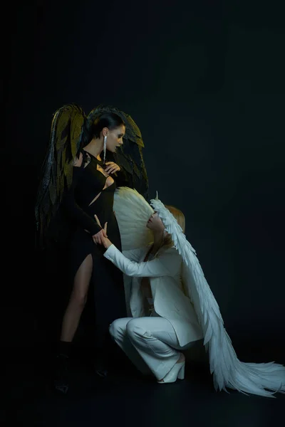 Himmlischer Engel an der Hand eines dunklen Dämons vor schwarzem Hintergrund, Frauen in Halloween-Kostümen — Stockfoto