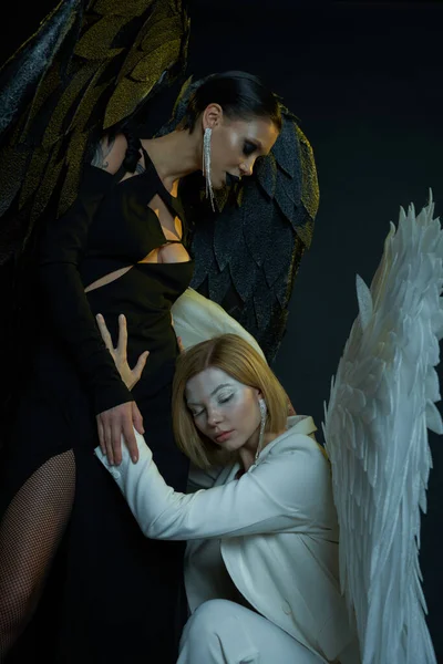 Women in Halloween costumes, angel embracing dark demon on black backdrop, biblical conflict concept — Stock Photo