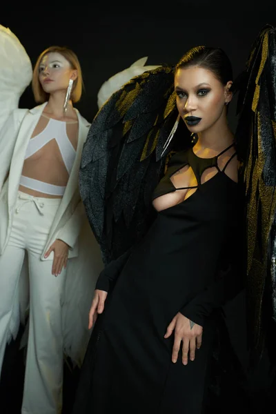 Donna demoniaca guardando macchina fotografica vicino angelo su sfondo nero, donne in costume di creature alate — Foto stock