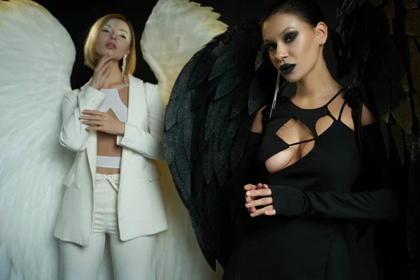 Mulher demoníaca olhando para a câmera perto de anjo no fundo preto, mulheres em trajes de criaturas aladas — Fotografia de Stock