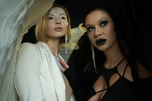 Mulheres em trajes de halloween de anjo e demônio olhando para longe em pano de fundo preto, guerra espiritual — Fotografia de Stock