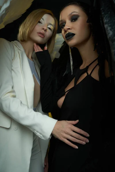 Mulheres em trajes de anjo e demônio olhando para a câmera no fundo preto, visão de baixo ângulo — Fotografia de Stock