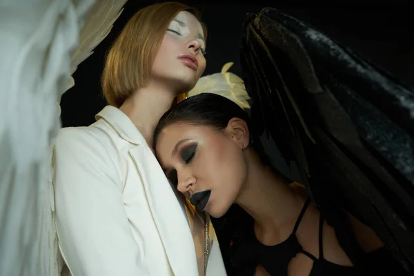Women in costumes of angel and demon with closed eyes on black, good vs evil biblical concept — Stock Photo