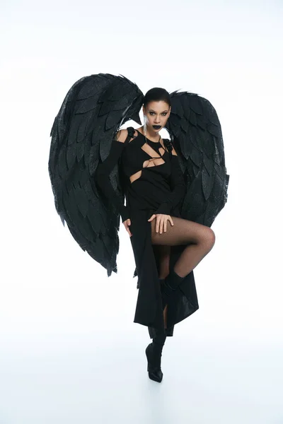 Piena lunghezza della donna in costume di angelo caduto nero con le ali guardando la fotocamera su sfondo bianco — Foto stock