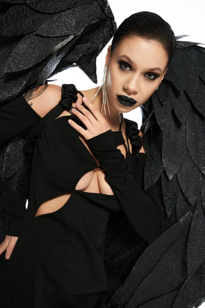Mujer encantadora en maquillaje oscuro y traje de criatura alada negro mirando a la cámara en blanco - foto de stock