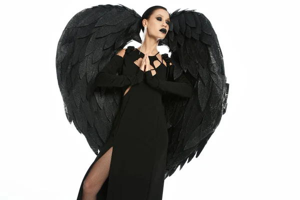 Mujer como demonio oscuro con alas negras rezando con los ojos cerrados en blanco, concepto de Halloween - foto de stock