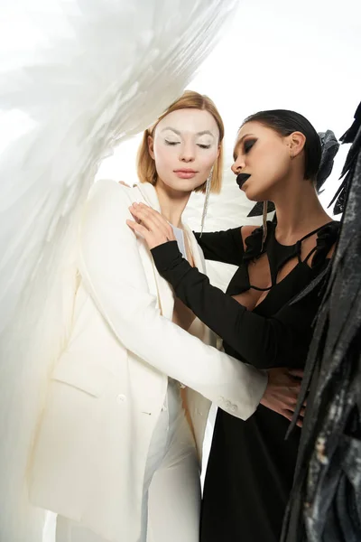 Women in costumes of dark and light winged creatures embracing on white, angel and demon concept — Stock Photo