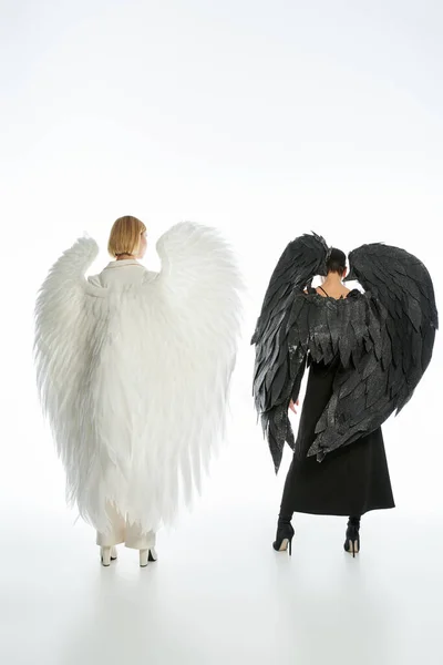 Задний вид женщин в костюмах дьявола и ангела с черными и светлыми крыльями на белом, полной длины — стоковое фото