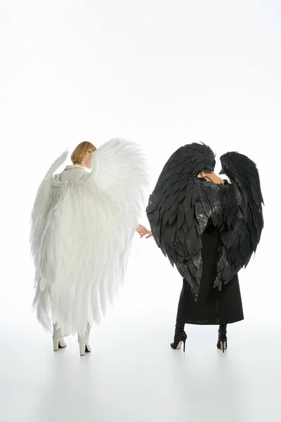 Задний вид женщин в костюмах дьявола и ангела с черными и светлыми крыльями, держащихся за руки на белом — стоковое фото