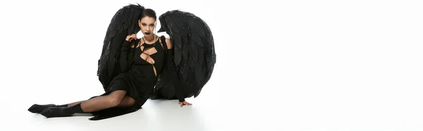 Сексуальная женщина в темном макияже и черном костюме крылатого демона сидит на белом, баннер — стоковое фото