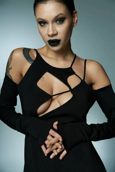 Séduisante femme tatouée avec un maquillage sombre portant une robe sexy noire et regardant la caméra sur gris — Photo de stock