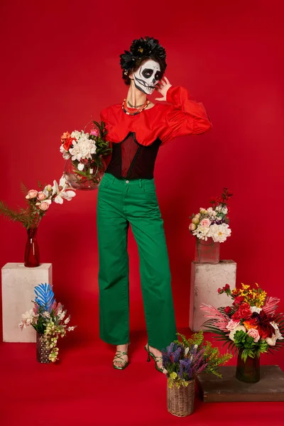 Woman in sugar skull makeup near traditional dia de los muertos altar with colorful flowers on red — Stock Photo
