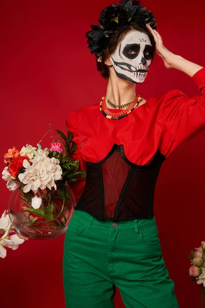Woman in skeleton makeup and festive attire holding vase with colorful flowers on red, Day of Dead — Stock Photo