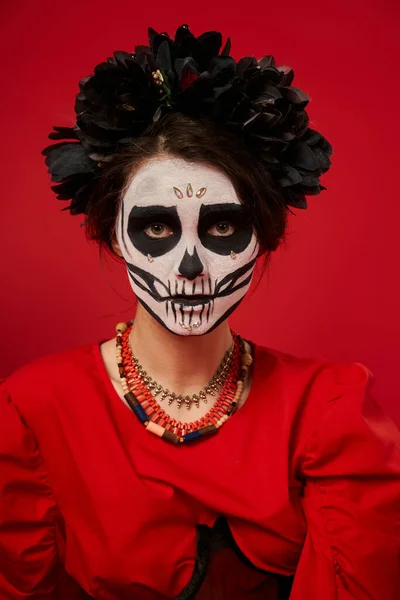 Femme en dia de los muertos maquillage et couronne noire avec des perles colorées regardant la caméra sur rouge — Photo de stock