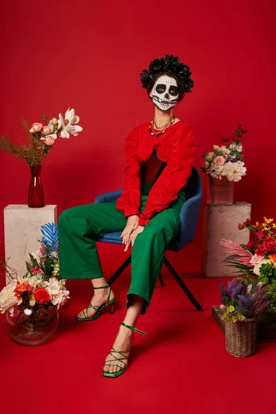 Femme dans dia de los muertos maquillage assis dans un fauteuil près de l'ofrenda traditionnel avec des fleurs sur rouge — Photo de stock