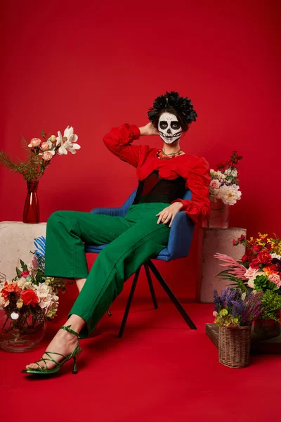 Woman in dia de los muertos makeup sitting in armchair near traditional altar with flowers on red — Stock Photo