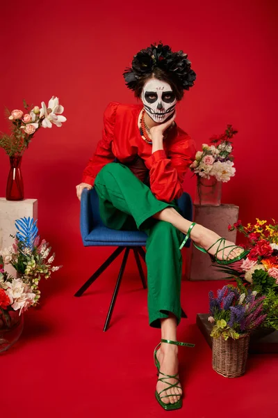 Femme dans le maquillage du crâne en regardant la caméra près de dia de los muertos ofrenda avec des fleurs sur rouge — Photo de stock