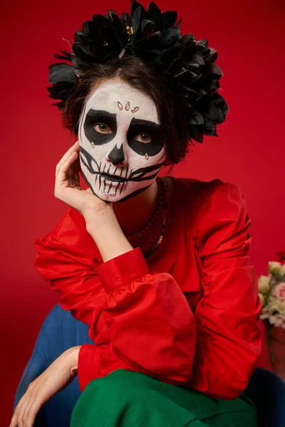 Retrato de mujer en maquillaje de cráneo y corona negra mirando a la cámara en rojo, tradición del Día de Muertos - foto de stock