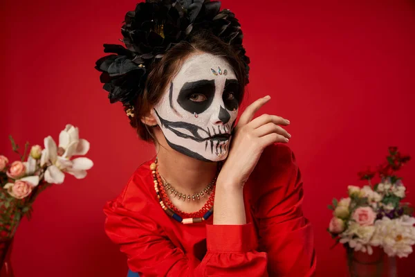 Mulher em assustador crânio maquiagem e coroa preta olhando para a câmera perto de flores no vermelho, retrato — Fotografia de Stock