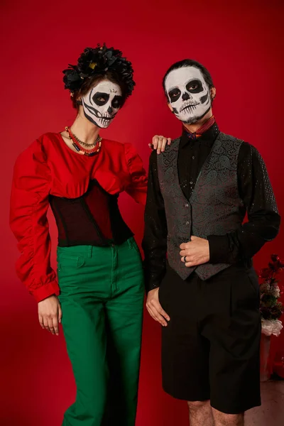 Elegant couple in dia de los muertos makeup and festive attire looking at camera near flowers on red — Stock Photo