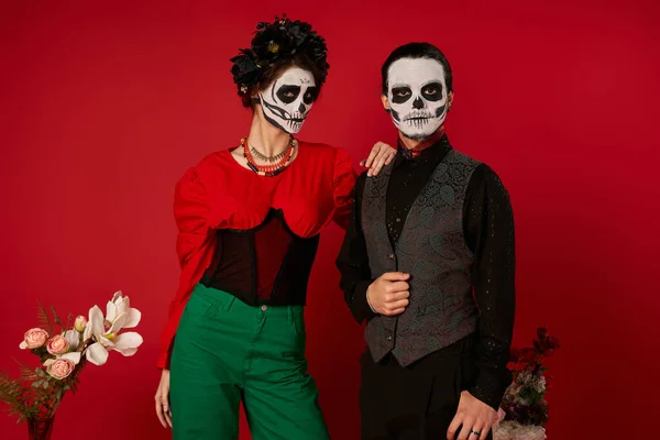 Stylish dia de los muertos couple in sugar skull makeup looking at camera near flowers on red — Stock Photo
